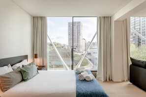 Ein Hotelzimmer mit einem großen Bett, einer Sitzbank mit Handtüchern, Blick auf eine Stadtlandschaft und einem modernen Bauwerk außen.
