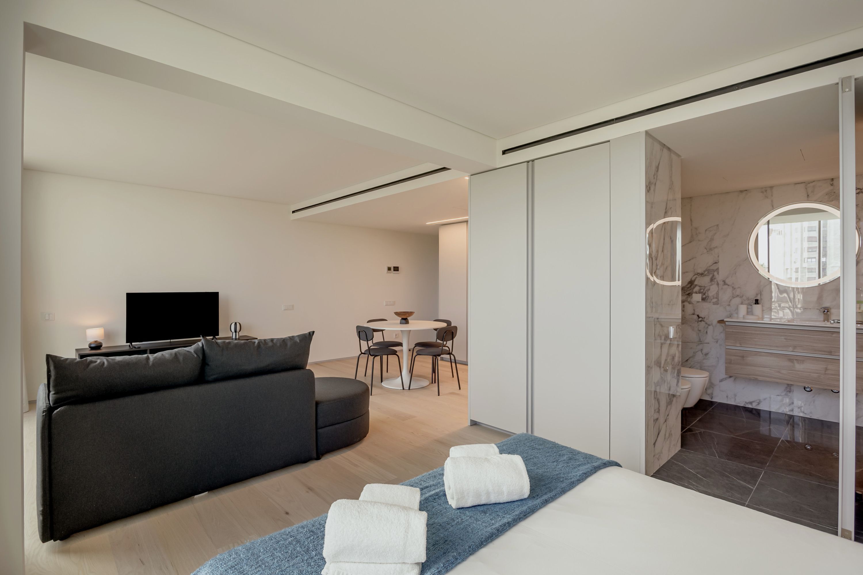 Una habitación de hotel moderna con sofá, cama con toallas, zona de comedor y baño con espejo.