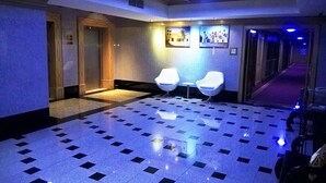 Un hall d’hôtel avec un sol à carreaux, deux chaises blanches, un comptoir de réception et un couloir avec des portes.