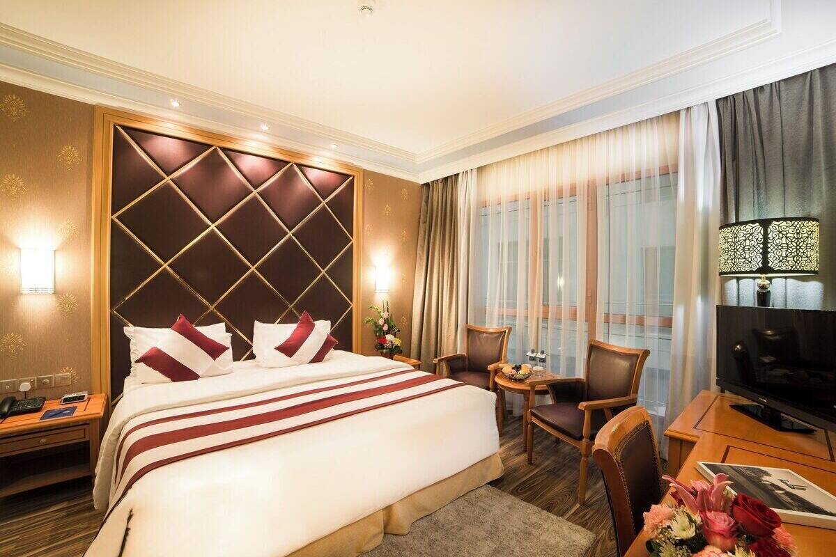 A hotel room with a large bed, a TV, a desk with a chair, and a small table with a lamp.