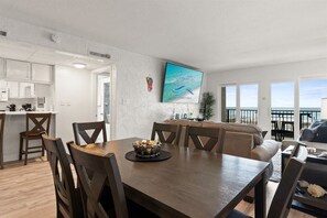 A dining area with a wooden table and chairs, a kitchen in the background, and a view of the ocean through large windows. - Beach View 2BR | Walk to Beach & Dining | Sleeps 6 (Panama City Beach)