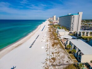 A beachside hotel with a long row of rooms along the shore, a clear blue sky, and a calm sea. - Beach View 2BR | Walk to Beach & Dining | Sleeps 6 (Panama City Beach)