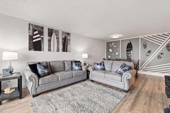 A living room with two grey sofas, a patterned rug, wall art, and a decorative wall panel. - Beach View 2BR | Walk to Beach & Dining | Sleeps 6 (Panama City Beach)