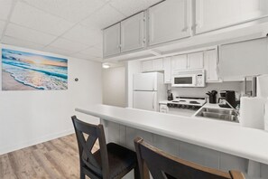 A modern kitchen with white cabinets, a microwave, a stove, and a sink. There is a painting of a beach scene on the wall. - Beach View 2BR | Walk to Beach & Dining | Sleeps 6 (Panama City Beach)