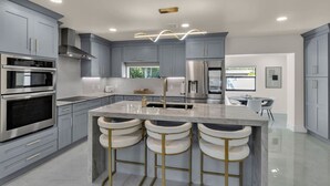 Una cocina moderna con gabinetes grises, electrodomésticos de acero inoxidable, una isla central con taburetes de bar e iluminación empotrada en el techo.