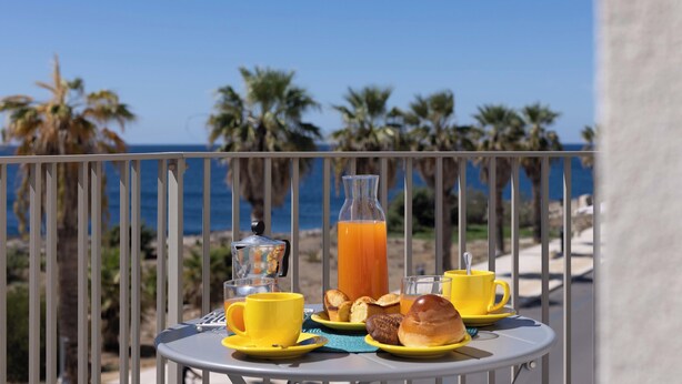 Een tafel met ontbijtartikelen en een glas sinaasappelsap op een balkon met uitzicht op zee.