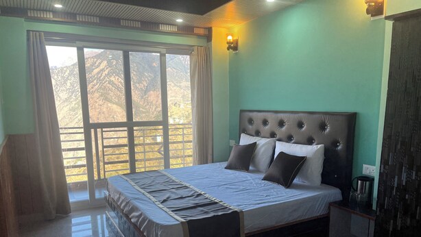 A bedroom with a large bed, a nightstand, a lamp, and a view of mountains through the window. - Your home away from home in mountains / 1-bedroom bed & breakfast (Rampur)