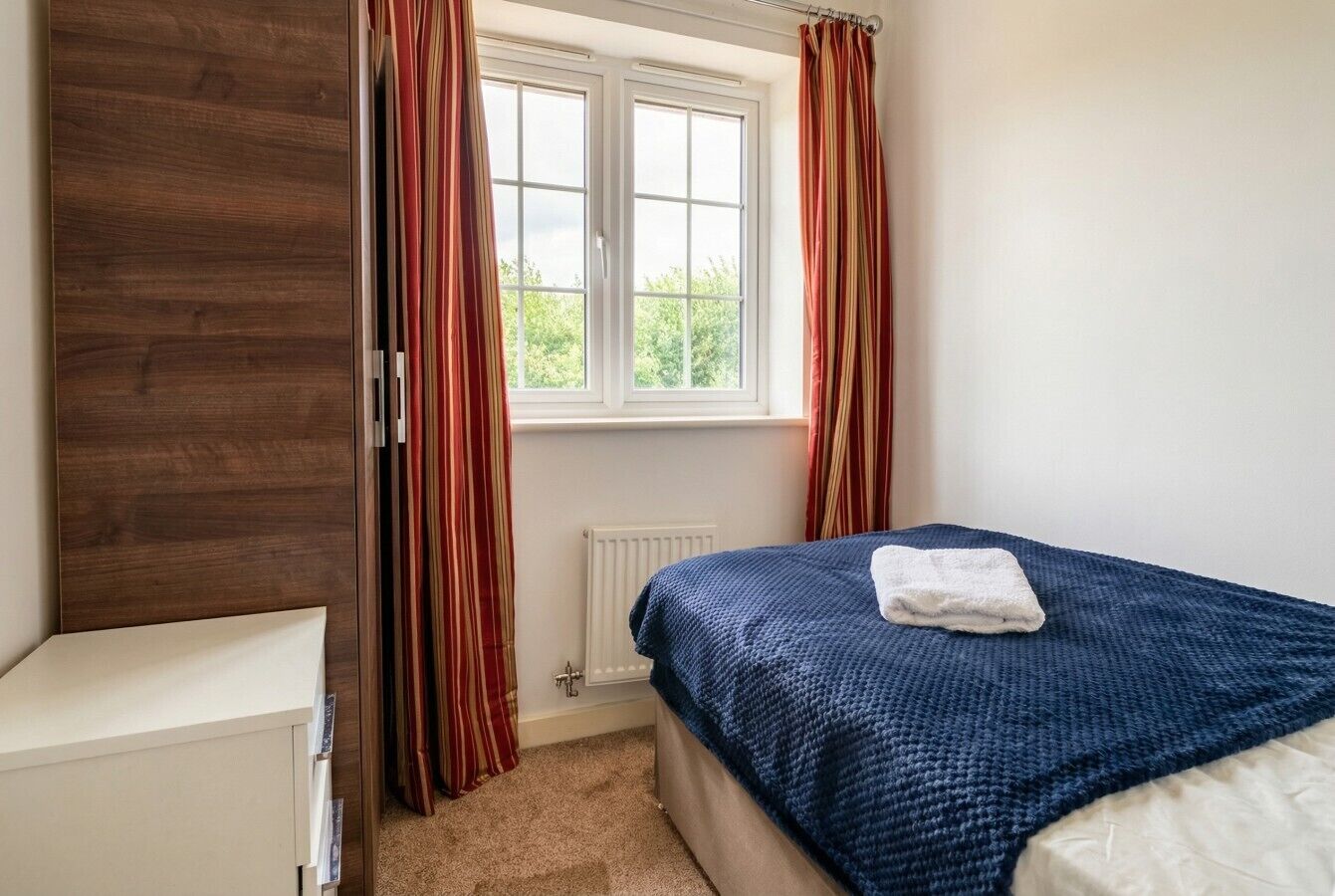 Une chambre à coucher avec un lit, une commode, une fenêtre avec des rideaux et un radiateur.