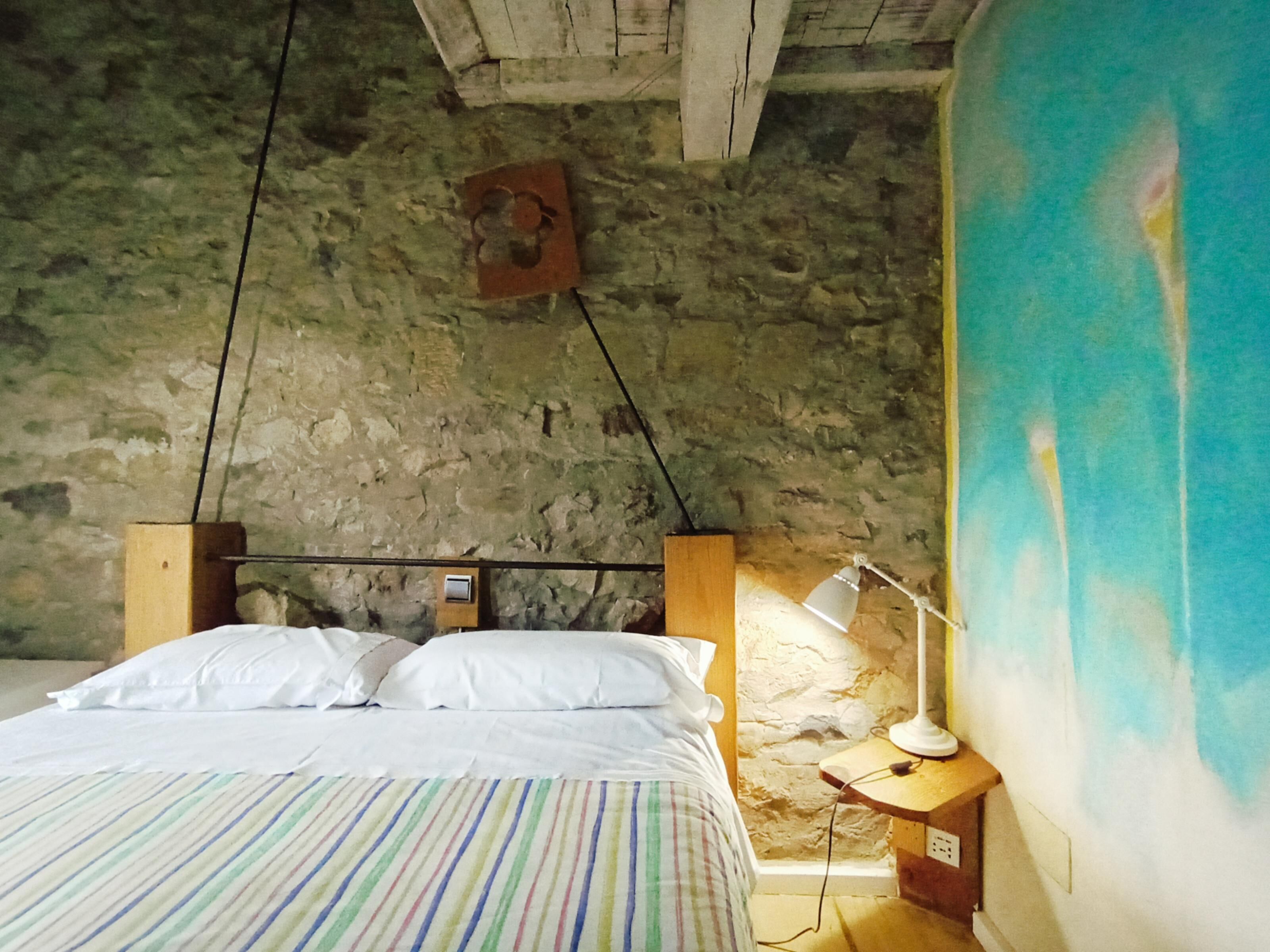 Una stanza con un letto, un comodino, una lampada, un muro in pietra e un grande quadro astratto.
