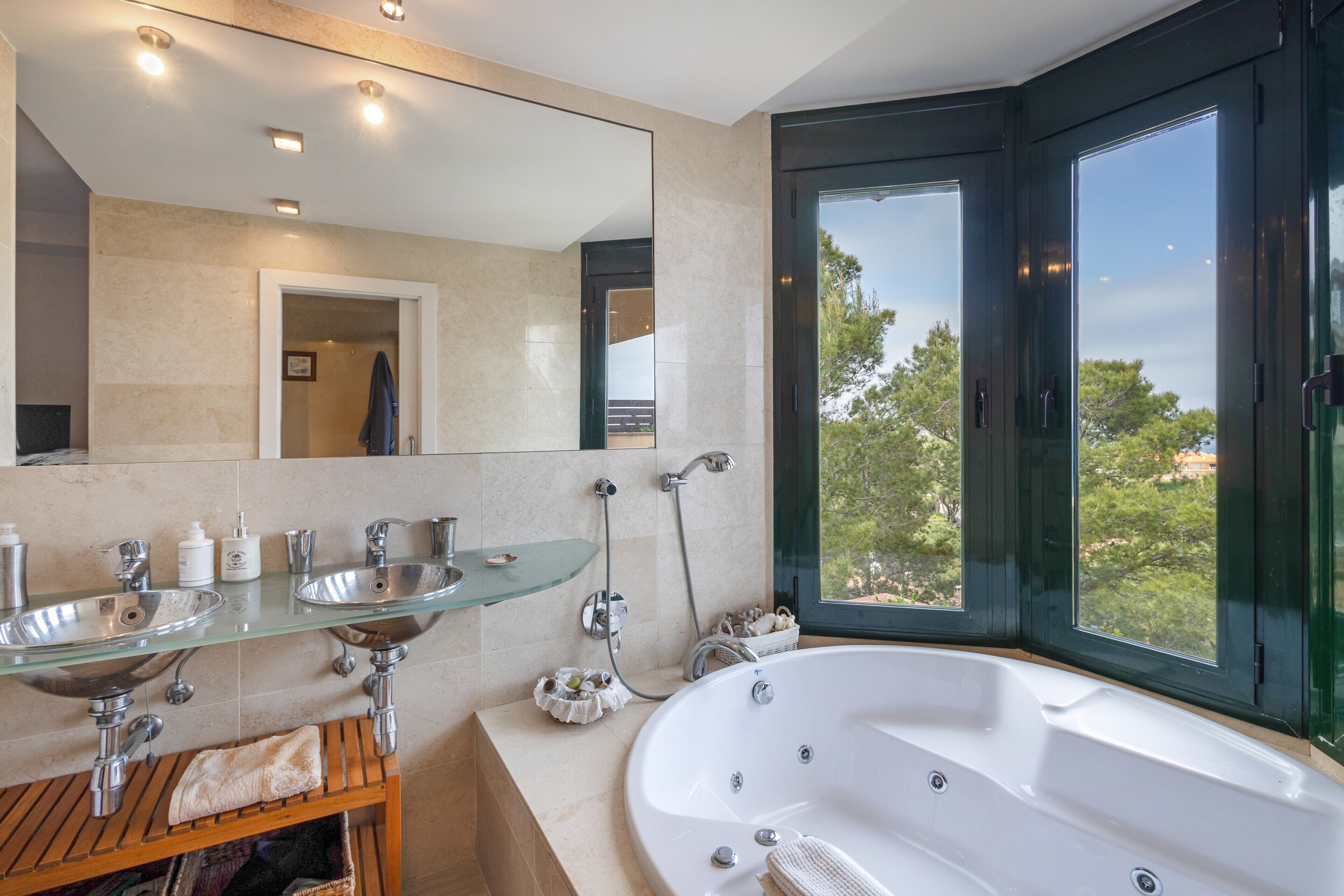 Ein modernes Badezimmer mit zwei Waschbecken aus Glas, einer großen Badewanne und Blick auf Bäume durch ein Fenster.