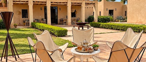 An outdoor patio with a table set for four, surrounded by lounge chairs, in front of a building with a covered entrance and a garden.