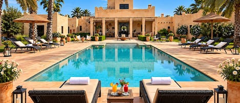 A poolside area with lounge chairs, a table with drinks, and a building with a distinctive architectural style in the background.