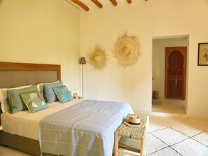 A bedroom with a bed, pillows, a woven chair, a lamp, and a door.