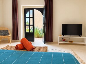 A bedroom with a bed, a television on a stand, a wicker chair, and a view of the outdoors through an open door.
