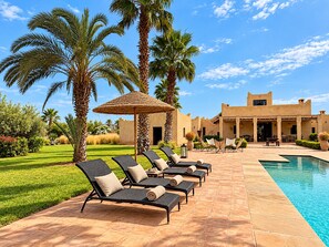 A poolside area with lounge chairs and a thatched umbrella, surrounded by palm trees and a building with a desert-style facade.