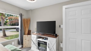 A modern living room with a TV on a wooden cabinet, a fireplace, and a view of a garden. - Cozy Beach Studio • Walk to the Sand (Pompano Beach)