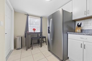 Eine kleine Küche mit einem Kühlschrank, einem Tisch für zwei Personen, einem Mülleimer und einem Fenster mit Vorhängen.