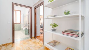 Um corredor com chão de azulejos, uma estante branca com livros e plantas, e uma porta que dá para o banheiro.