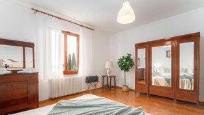 Quarto com cama, penteadeira, espelho, radiador, janela com cortinas, luminária, planta e guarda-roupa.