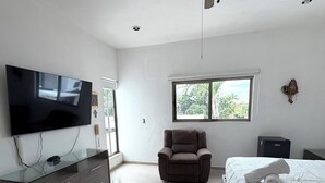 Un dormitorio con cama, televisor, sillón, ventilador de techo y ventana.