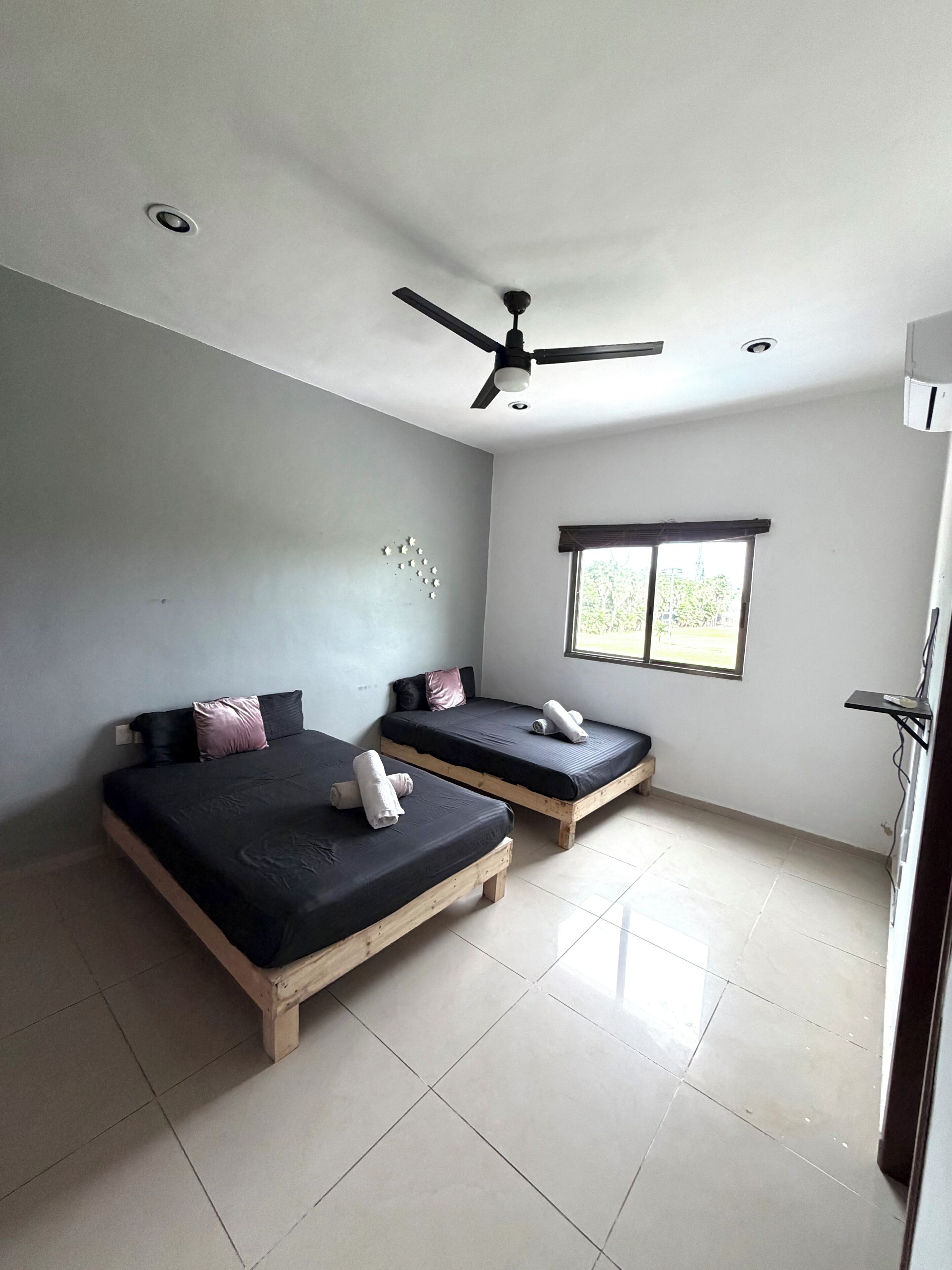 A room with two wooden beds, a ceiling fan, a window with blinds, and a tiled floor.