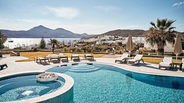 Un espace piscine doté de chaises longues et d’un jacuzzi, offrant une vue sur une ville côtière et des montagnes.