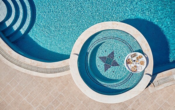Une piscine circulaire ornée d’une mosaïque en forme d’étoile, une terrasse carrelée et un transat avec un plateau de collations.
