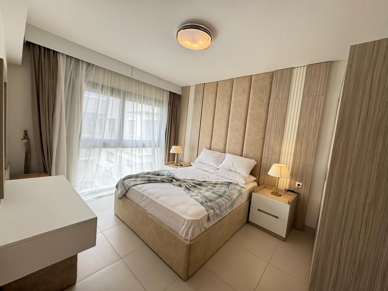 A modern bedroom with a bed, bedside tables, a nightstand, a desk, and a large window with curtains.