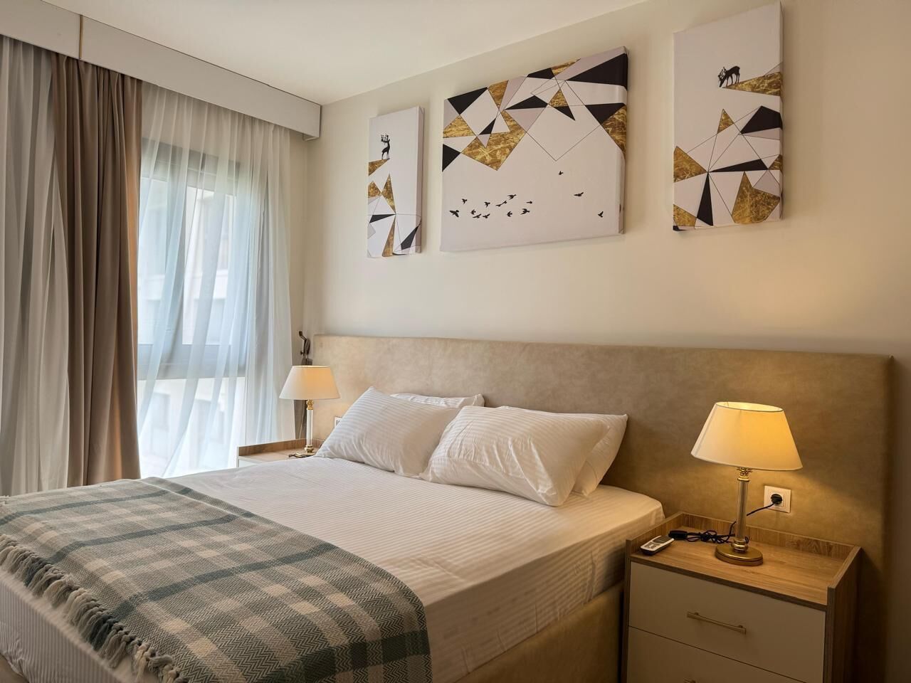 Una habitación de hotel con una cama, dos mesitas de noche, una lámpara, un edredón y tres obras de arte abstracto en la pared.