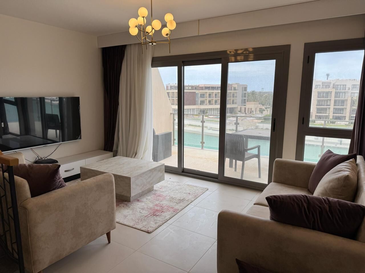 A modern living room with a flat-screen TV, two sofas, a coffee table, and large sliding glass doors leading to a pool area.