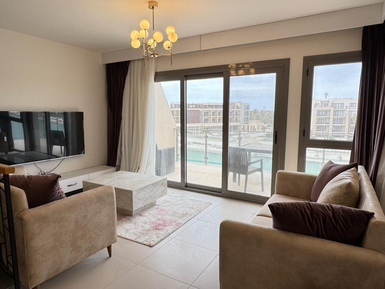 A modern living room with a flat-screen TV, two sofas, a coffee table, and a large sliding glass door leading to a balcony with a pool view.