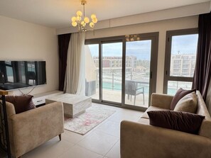 Ein modernes Wohnzimmer mit einem Flachbildfernseher, zwei Sofas, einem Couchtisch und einer großen Glaswand, die auf einen Balkon mit Blick auf einen Pool führt.