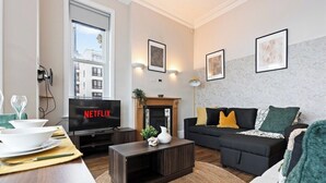 Ein Wohnzimmer mit Fernseher, auf dem 'NETFLIX' läuft, einem Kamin, einer Couch und einem Couchtisch.