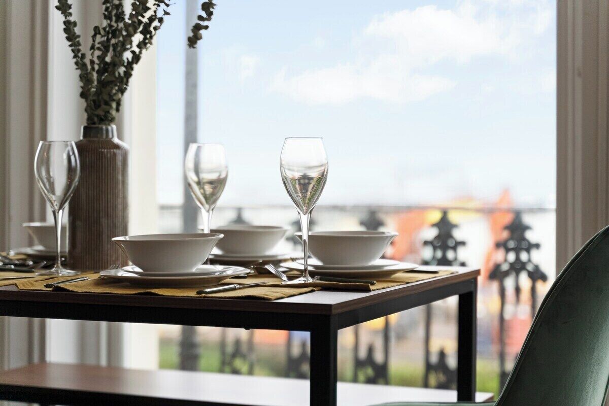 A dining table set with white bowls and wine glasses, a vase with greenery, and a view of a cityscape through a window.