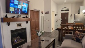 Una sala de estar con chimenea, televisor montado en la pared, comedor con mesa y sillas, y un sofá con cojín.