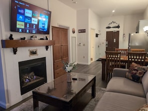 A living room with a fireplace, a television mounted on the wall, a dining area with a table and chairs, and a sofa with a cushion. - Amazing 1st floor Ski in/out Unit @ Creek (Vernon Township)