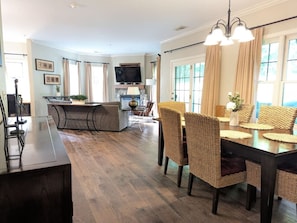 A dining area with a wooden table and wicker chairs, a kitchen with a TV, and a living area with a sofa and a fireplace.