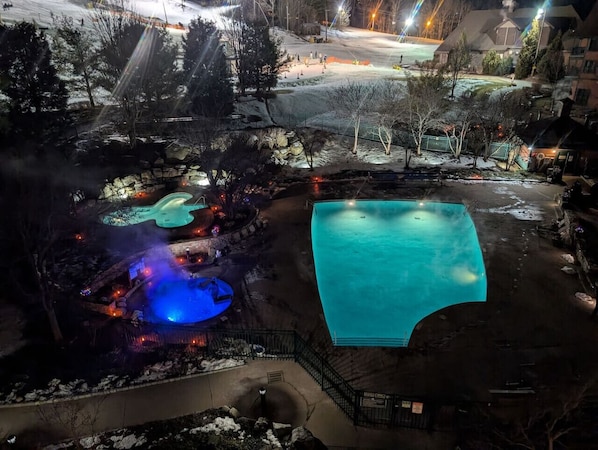 Une grande piscine extérieure entourée d’un espace aménagé, comprenant une piscine plus petite et un espace avec un jacuzzi, le tout agrémenté d’un éclairage.