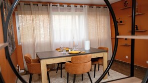 A dining area with a table set for four, chairs, a window with curtains, and a wall with shelves.