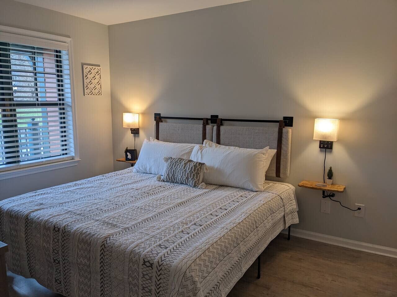 A bedroom with a bed, two bedside lamps, a window with blinds, and a wall-mounted shelf with a plant.