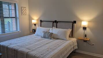 A bedroom with a bed, two bedside lamps, a window with blinds, and a wall-mounted shelf with a plant.