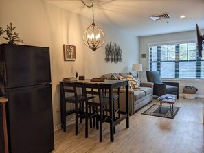 Ein modernes Wohnzimmer mit Essbereich, einem schwarzen Kühlschrank, einer Chandelierleuchtre und einem Fenster mit Jalousien.