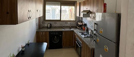 A kitchen with wooden cabinets, a black countertop, a stainless steel refrigerator, and a window with a view of buildings.