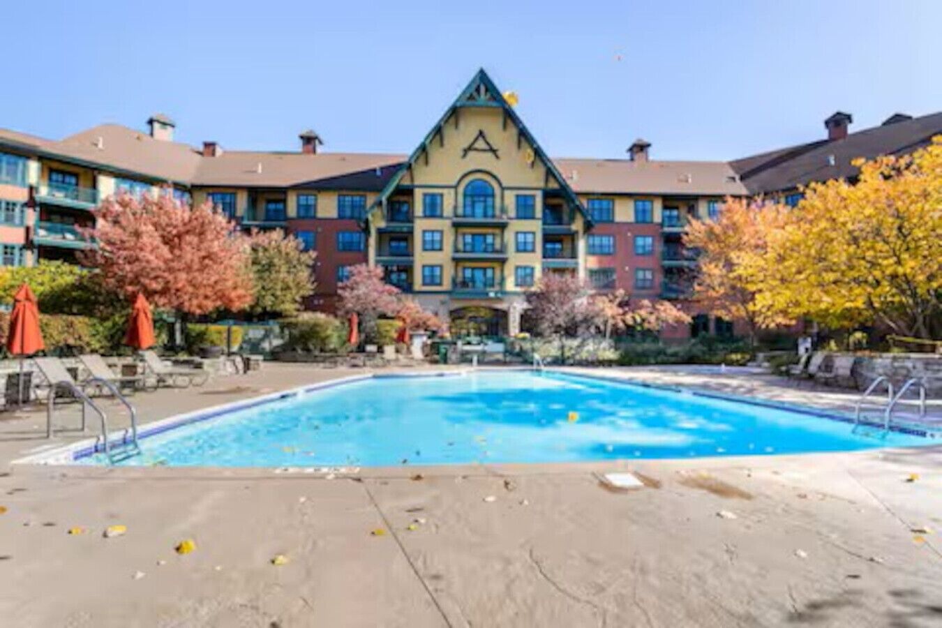 A hotel with a swimming pool, surrounded by autumn-colored trees and a multi-story building with a distinctive triangular roof.