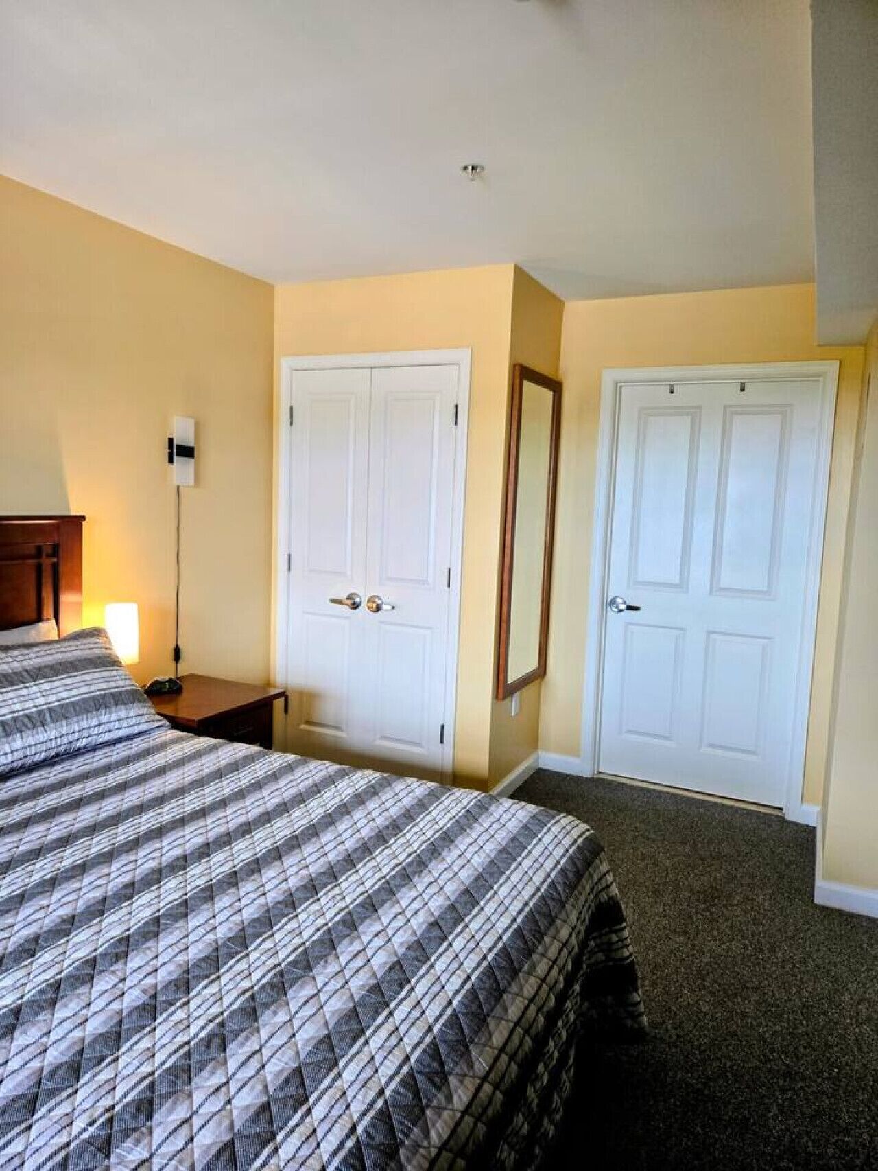 A hotel room with a bed, two white doors, a nightstand, a lamp, and a mirror.