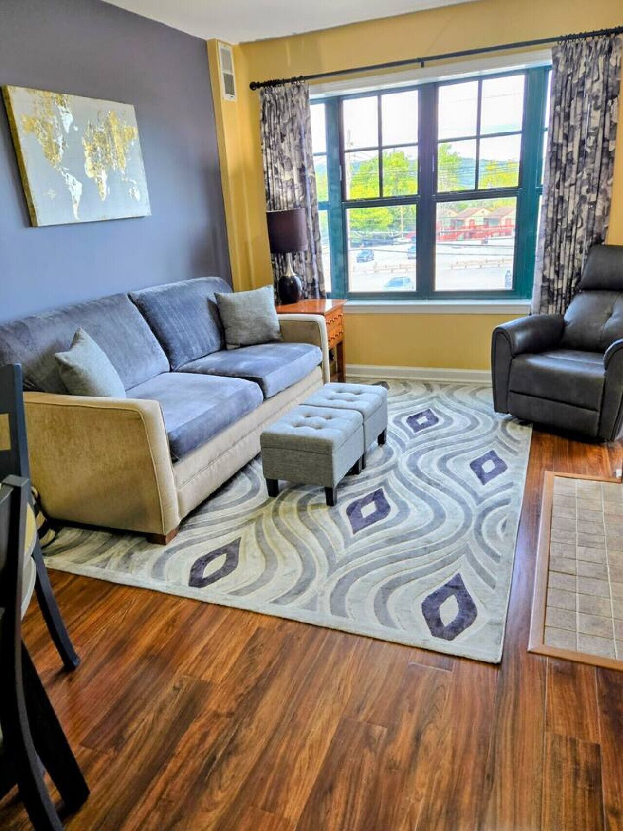 A living room with a sofa, ottoman, and chair, a patterned rug, and a window with curtains.