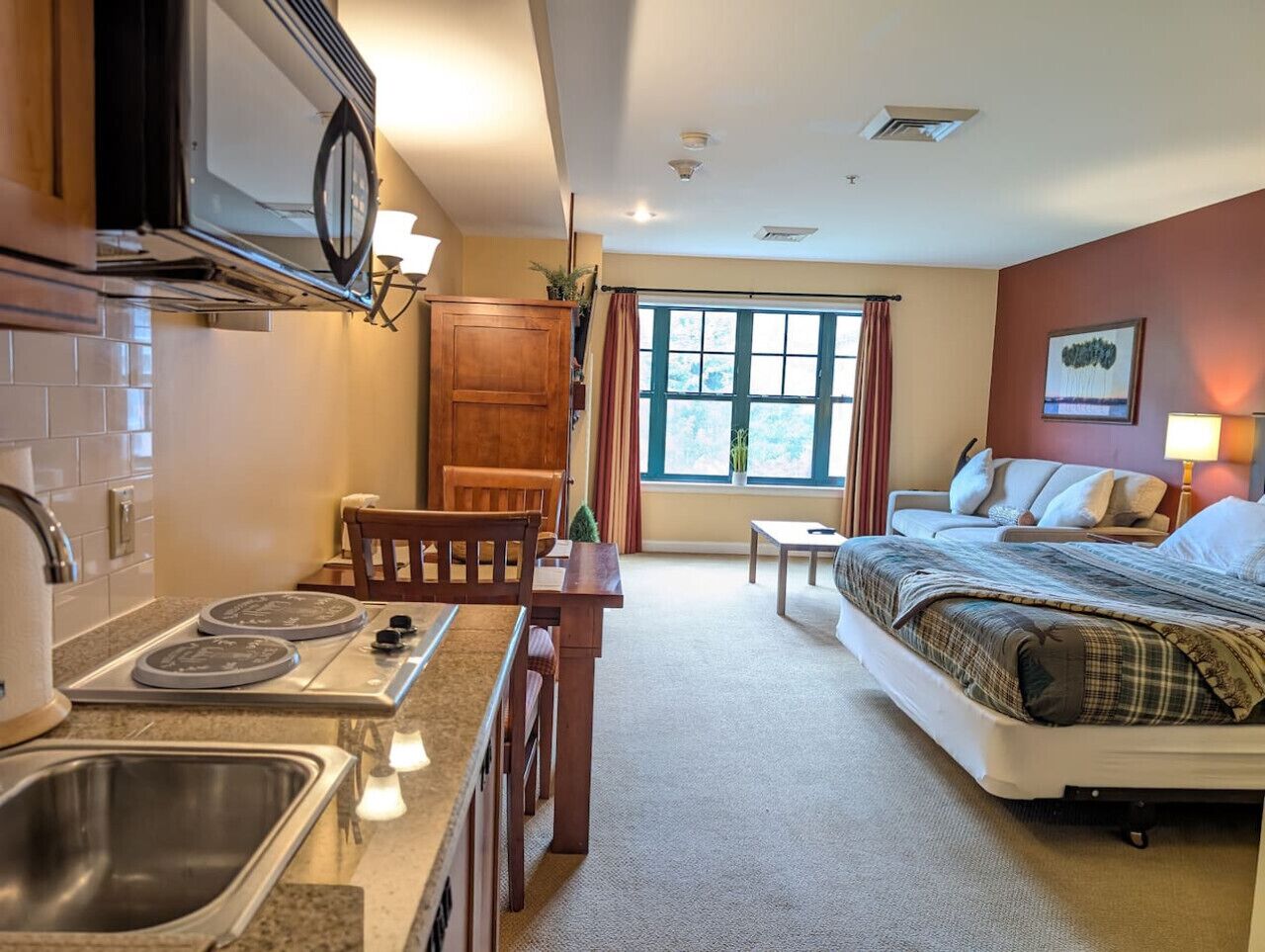 Une chambre d’hôtel compacte comprenant un coin cuisine, un lit, une petite table et une fenêtre avec des rideaux.