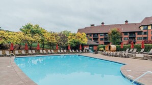 Ein Schwimmbecken mit Liegestühlen und Sonnenschirmen, umgeben von Bäumen und einem Gebäude mit mehreren Fenstern.