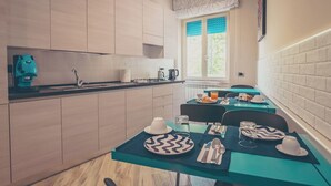 A modern kitchen with a built-in stove, a coffee machine, and a window with blue curtains. There are two dining tables with blue tablecloths, each set with plates, cutlery, and glasses. - Cozy B&B,right in the center & within walking distance of beautiful sea of Gaeta (Gaeta)