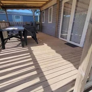 A covered patio area with a wooden deck, a table with chairs, and a sliding glass door. - Family mobile home Camping 5* Le Marisol (Torreilles)
